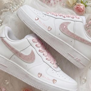 May include: A pair of white sneakers featuring pink glitter swooshes, pink satin laces, and small pink heart accents. The side of each shoe is customized with the text Mrs. Clark in pink script lettering, set against a white leather base.
