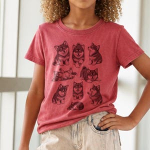 Pode incluir: Uma t-shirt de manga curta cor carmesim e uma t-shirt de manga curta cor ganga lavada, ambas com uma impressão gráfica preta de vários cães Shiba Inu em várias poses. O texto inclui 20+ Colors, 3 tee types, Crimson e Washed Denim.