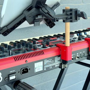 May include: A black plastic tablet mount attached to a red keyboard via a wooden dowel and red clamps. The mount features an adjustable arm and a cradle holding a white tablet. The keyboard has black knobs, buttons, and a power switch visible.