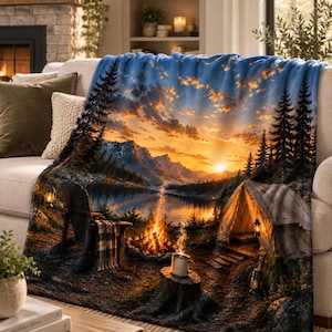 May include: A plush throw blanket featuring a vibrant landscape print of a mountain lake at sunset. The scene includes a glowing campfire, a camping tent, a wooden chair with a plaid blanket, and a steaming mug resting on a tree stump.
