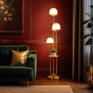 May include: A tall floor lamp with a gold-toned metal frame featuring three stacked tiers, each holding a spherical white glass light globe. A small circular shelf is integrated into the structure, supporting a potted green plant.