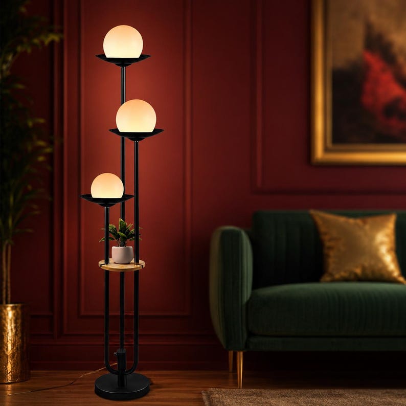 May include: A black floor lamp featuring three vertical poles of varying heights, each topped with a white spherical glass shade. A small circular wooden shelf is integrated into the structure, holding a small green potted plant.