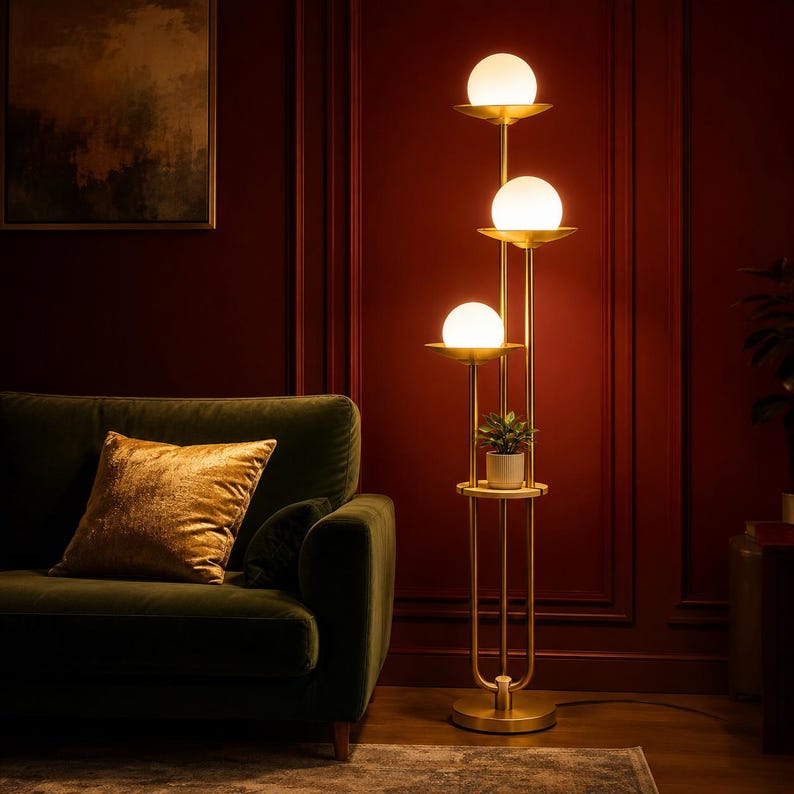 May include: A floor lamp with a gold-toned metal frame featuring three vertical poles of varying heights, each topped with a spherical white glass shade. A circular shelf is integrated into the frame, holding a small potted green plant.