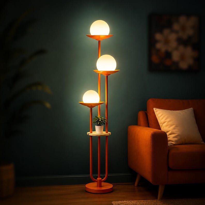 May include: A tall orange floor lamp featuring three vertical poles of varying heights, each topped with a glowing white spherical glass shade. A small circular wooden shelf is integrated into the structure, holding a small potted green plant.