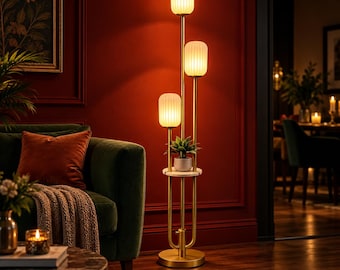 May include: A floor lamp with a gold-toned metal frame featuring three vertical poles of varying heights, each topped with a ribbed white glass shade. A circular white marble shelf is positioned in the center, holding a small potted green plant.