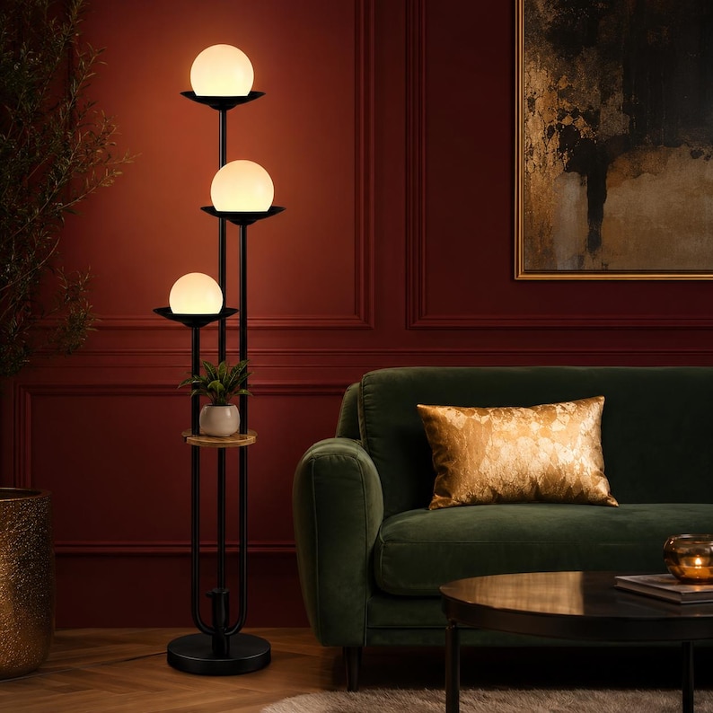 May include: A tall floor lamp with a black metal frame featuring three stacked white spherical glass shades. The lamp includes a small circular wooden shelf positioned in the middle, holding a small potted green plant with white leaves.