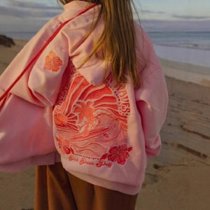 May include: A light pink hooded sweatshirt featuring a red graphic on the back depicting a palm tree, a surfboard, ocean waves, and hibiscus flowers. The text reads "EVERYTHING IS IN WAVES" and "Pink Palm Puff" in red lettering.
