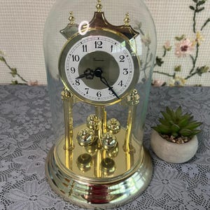 May include: A gold-toned anniversary clock with a circular white face, black Arabic numerals, and ornate black hands, enclosed within a clear glass dome. The clock features a rotating pendulum with four gold spheres on a circular base.
