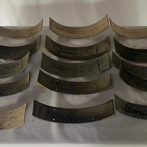 May include: A collection of fourteen curved, semi-circular brake shoe linings arranged in rows. Each piece features a textured, tan or dark grey surface with small pre-drilled holes and the Ford logo printed in black ink along with various part numbers.
