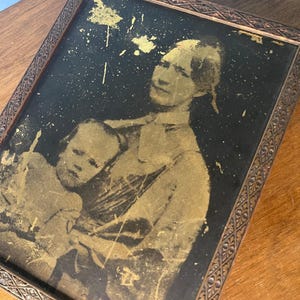 May include: A rectangular framed portrait featuring a gold-toned image of an adult holding a child against a black background. The print shows signs of wear with gold flecks scattered across the surface. The frame has a dark brown ornate pattern.
