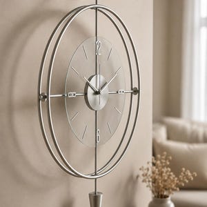 Op de afbeelding: Een wandklok met een cirkelvormig zilverkleurig metalen frame met twee concentrische ringen. Een transparante wijzerplaat toont zilveren uurmarkeringen en de cijfers 12 en 6. Een centrale zilveren schijf bevat de wijzers en een slinger.