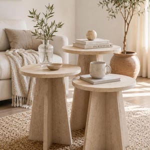 Può includere: Un set di tre tavolini rotondi realizzati in pietra travertino color beige chiaro, ciascuno caratterizzato da un piano circolare piatto sostenuto da una robusta base a piedistallo con più pannelli. I tavoli variano in altezza, creando una disposizione a gradini.