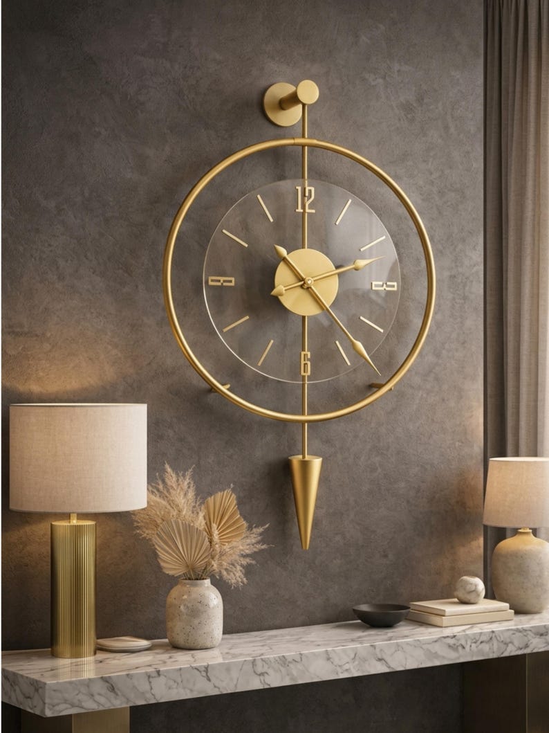 May include: A wall clock featuring a circular gold frame and a transparent center dial. The clock has gold hands, gold hour markers, and the numbers 12 and 6 in gold. A vertical gold rod extends from the top to a pointed gold pendulum at the bottom.