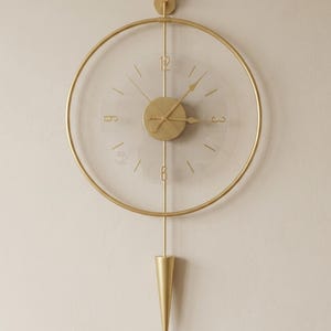 May include: A circular wall clock featuring a gold-toned metal frame and a transparent face. The clock includes gold-toned hands, numerals at the 12, 3, 6, and 9 positions, and a central gold disc. A vertical gold rod extends to a cone-shaped pendulum.