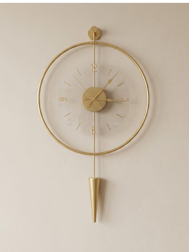 May include: A circular wall clock featuring a gold-toned metal frame and a transparent face. The clock includes gold-toned hands, numerals at the 12, 3, 6, and 9 positions, and a central gold disc. A vertical gold rod extends to a cone-shaped pendulum.