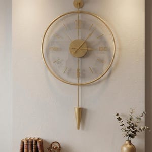 May include: A circular wall clock with a gold-toned metal frame and a transparent face. It features gold-colored hands, a central circular hub, and vertical bars extending to a pointed, cone-shaped pendulum at the bottom. The numbers 12 and 6 are displayed in a gold font.
