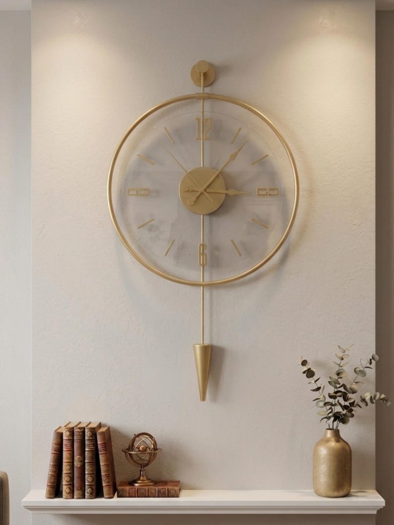 May include: A circular wall clock with a gold-toned metal frame and a transparent face. It features gold-colored hands, a central circular hub, and vertical bars extending to a pointed, cone-shaped pendulum at the bottom. The numbers 12 and 6 are displayed in a gold font.