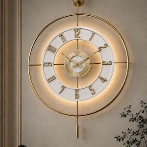 Op de afbeelding: Een grote ronde wandklok met een gouden metalen frame, een witte wijzerplaat met verhoogde gouden cijfers en een gouden centrum. Het ontwerp bevat geïntegreerde achtergrondverlichting, dunne gouden wijzers en een verticale gouden slinger.