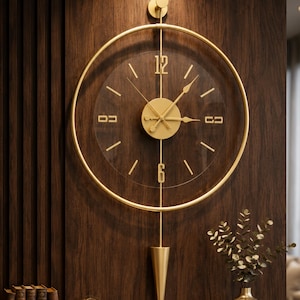 Può includere: Un orologio da parete circolare con cornice in metallo dorato e quadrante trasparente. Presenta lancette dorate, una piastra centrale circolare e i numeri 12 e 6. Un'asta verticale si estende verso un pendolo a forma di cono sul fondo.
