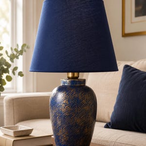 May include: A table lamp featuring a navy blue fabric drum shade and a rounded ceramic base. The base has a textured navy blue finish with distressed gold-colored cross-hatch detailing and a metallic gold-toned neck piece at the top.