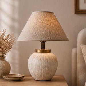 Może przedstawiać: Lampa stołowa z okrągłą, teksturowaną białą ceramiczną podstawą z pionowymi brązowymi smugami, mosiężną metalową szyjką i stożkowym kloszem z jasnobeżowej tkaniny o widocznej splocie, emitująca miękkie, ciepłe światło.