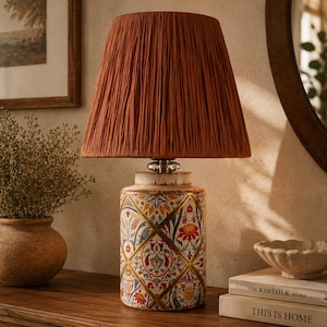 May include: A table lamp with a pleated, rust-colored fabric shade and a cylindrical ceramic base featuring a colorful, diamond-patterned floral design with gold accents. The lamp sits on a wooden surface next to two stacked books.