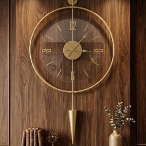 May include: A large circular wall clock with a gold-toned metal frame and a transparent face. The clock features gold-colored hands, numerals at the 12 and 6 positions, and a vertical gold pendulum with a cone-shaped weight hanging below the dial.