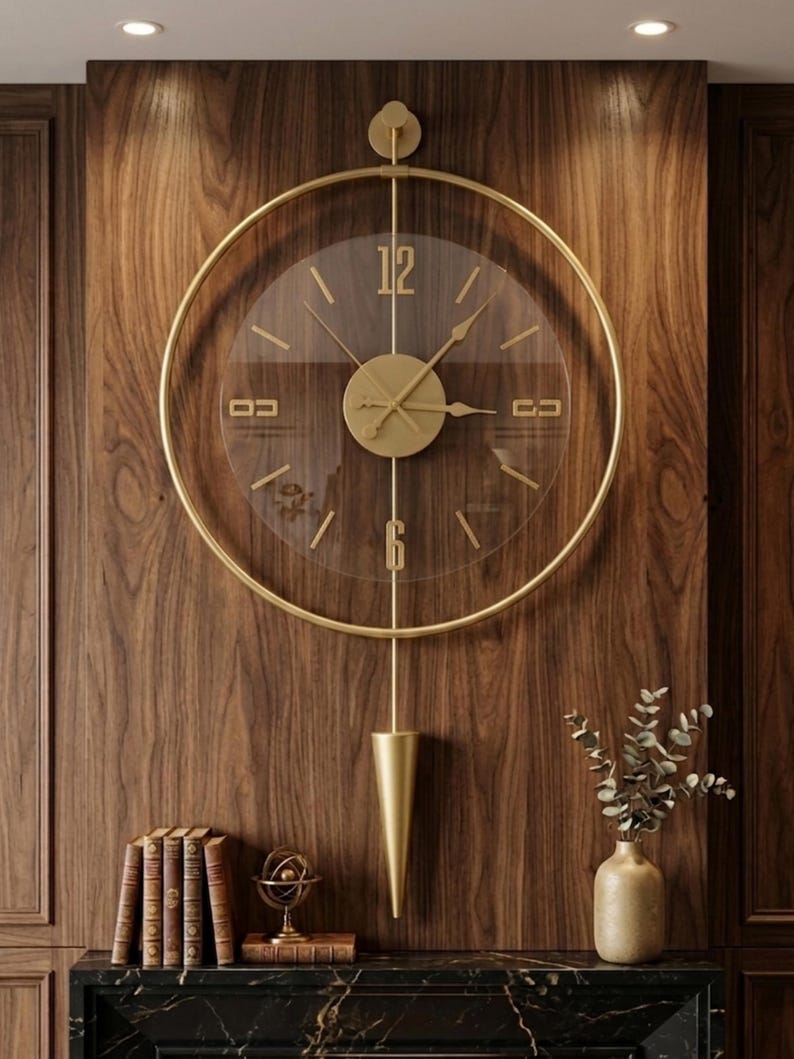 May include: A large circular wall clock with a gold-toned metal frame and a transparent face. The clock features gold-colored hands, numerals at the 12 and 6 positions, and a vertical gold pendulum with a cone-shaped weight hanging below the dial.