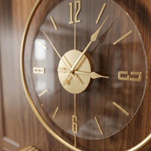 May include: A circular wall clock featuring a gold-toned metal frame and a transparent face. The clock has gold-colored hour markers, including the numerals 12 and 6, and matching gold hands. A central gold disc houses the clock mechanism.