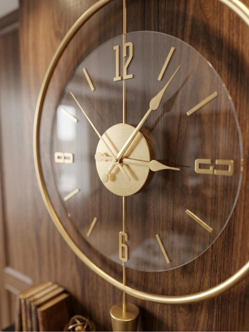 May include: A circular wall clock featuring a gold-toned metal frame and a transparent face. The clock has gold-colored hour markers, including the numerals 12 and 6, and matching gold hands. A central gold disc houses the clock mechanism.