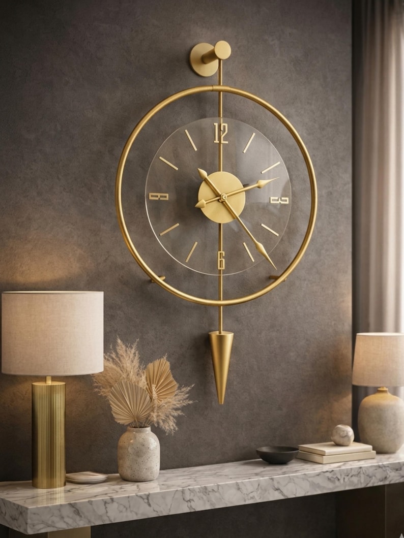 May include: A wall clock featuring a circular gold metal frame and a transparent center dial. The clock has gold-colored hands and numerals at the 12, 3, 6, and 9 positions. A vertical gold rod extends from the top mount to a pointed gold pendulum.
