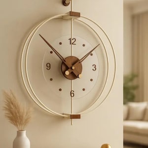 May include: A wall clock featuring a clear circular face with dark brown numerals at 3, 6, 9, and 12 o'clock. The clock has a dark brown wooden centre, thin dark brown hands, and is framed by two large, thin, off-white circular metal rings.