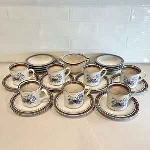 May include: A ceramic dinnerware set featuring six mugs with matching saucers, two small bowls, and a creamer pitcher. Each piece is off-white with brown rim accents and a blue crest graphic depicting a shield, arrows, and a ribbon banner design.
