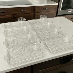 May include: A set of five clear glass rectangular serving trays and five matching glass teacups. Each tray features a textured pattern of concentric circles and small raised dots, with integrated handles on the short ends of the trays.