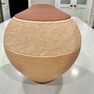 May include: A spherical ceramic vase featuring a smooth, matte terracotta-colored top section and a textured lower body. The middle portion displays a pattern of fine, diagonal incised lines carved into the light beige clay surface.