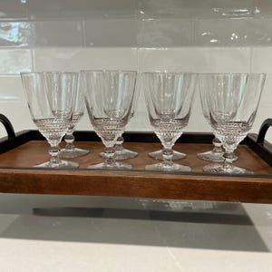 May include: A set of six clear glass goblets with a textured, beaded pattern at the base of the bowl and on the stem. The glasses are arranged on a rectangular wooden serving tray with black metal handles and a dark brown finish.