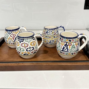 May include: A set of four ceramic mugs with rounded bodies and handles, each featuring intricate hand-painted geometric and floral patterns in vibrant shades of blue, yellow, green, and red against a cream-colored background with dark blue rims.