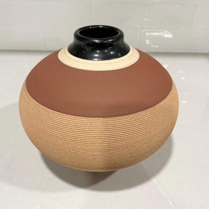 May include: A spherical ceramic vase featuring a smooth, matte terracotta-colored upper section and a textured, ribbed lower section in a light tan hue. The neck is finished with a glossy black glaze and a thin, cream-colored band.