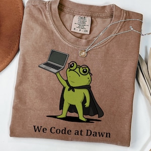 May include: A brown short-sleeve t-shirt featuring a graphic of a green frog wearing glasses and a black cape, holding a laptop. The text "We Code at Dawn" is printed below the frog. The shirt has a Comfort Colors brand label at the neck.
