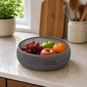 May include: A round, grey, open-weave plastic fruit bowl containing a cluster of red grapes, a green apple, a red apple, and an orange. The bowl features a repeating geometric lattice pattern and sits on a light-colored countertop surface.