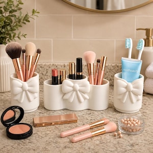 May include: A set of three white ceramic organizers featuring embossed bow details. Two cylindrical holders contain makeup brushes and toothbrushes with a blue tube labeled roothoastg, while the central oval holder stores lipsticks and brushes.