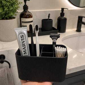 May include: A black rectangular bathroom organizer with vertical ribbed texture, containing a tube of Marvis toothpaste, one black toothbrush, one white toothbrush, a razor, and a small bundle of cotton swabs in separate compartments.