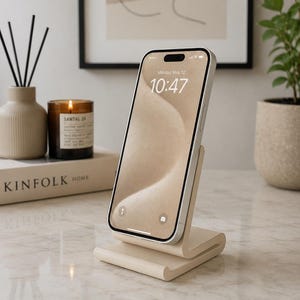May include: A light beige smartphone stand with a curved, minimalist design holding a smartphone in an upright position. The phone screen displays a beige sand-colored background with the time 10:47 and the date Monday, May 12 clearly visible.
