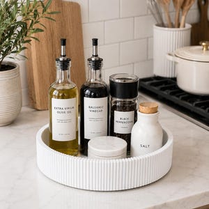 May include: A white circular ribbed tray containing two glass bottles labeled Extra Virgin Olive Oil and Balsamic Vinegar, a black pepper grinder, a small white marble container, and a white ceramic salt jar with a cork lid.