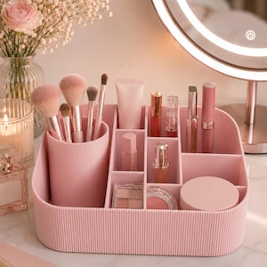 May include: A light pink plastic makeup organizer with a ribbed exterior texture, featuring a cylindrical brush holder and multiple rectangular compartments containing various cosmetic items like brushes, lip gloss tubes, and compacts.