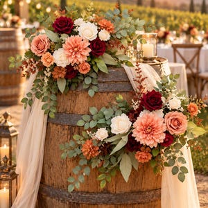 May include: A rustic wooden barrel decorated with two large floral arrangements featuring deep red, peach, and white flowers, including dahlias and roses, accented with green eucalyptus leaves and draped with sheer cream fabric, set in an outdoor setting.