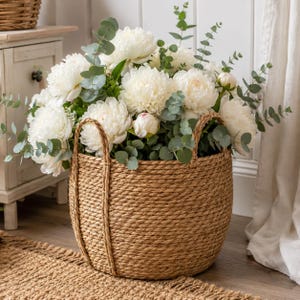 May include: A round, woven seagrass basket with two sturdy handles, filled with a large arrangement of white peony flowers and green eucalyptus stems. The basket features a thick, coiled rope texture and a natural tan color throughout.