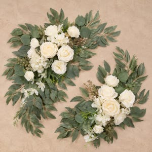 May include: Two floral arrangements featuring cream-colored roses and white hydrangea blossoms accented with various green eucalyptus leaves and small sprigs of filler greenery. The flowers are arranged in a cascading, elongated design.