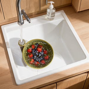 Op de afbeelding: Een witte vierkante gootsteen met een chromen kraan die water in een groene kom vol verse rode aardbeien en donkere bosbessen laat lopen. Een doorzichtige zeepdispenser staat op de rand van de gootsteen, die in een lichthouten aanrechtblad is geplaatst.