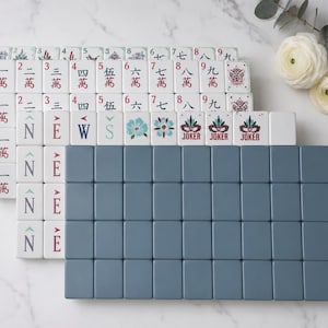 May include: A set of rectangular mahjong tiles arranged in rows. The tiles feature white faces with red, blue, and green printed Chinese characters, floral motifs, and the word JOKER. A large block of solid slate blue tiles is positioned in the front.